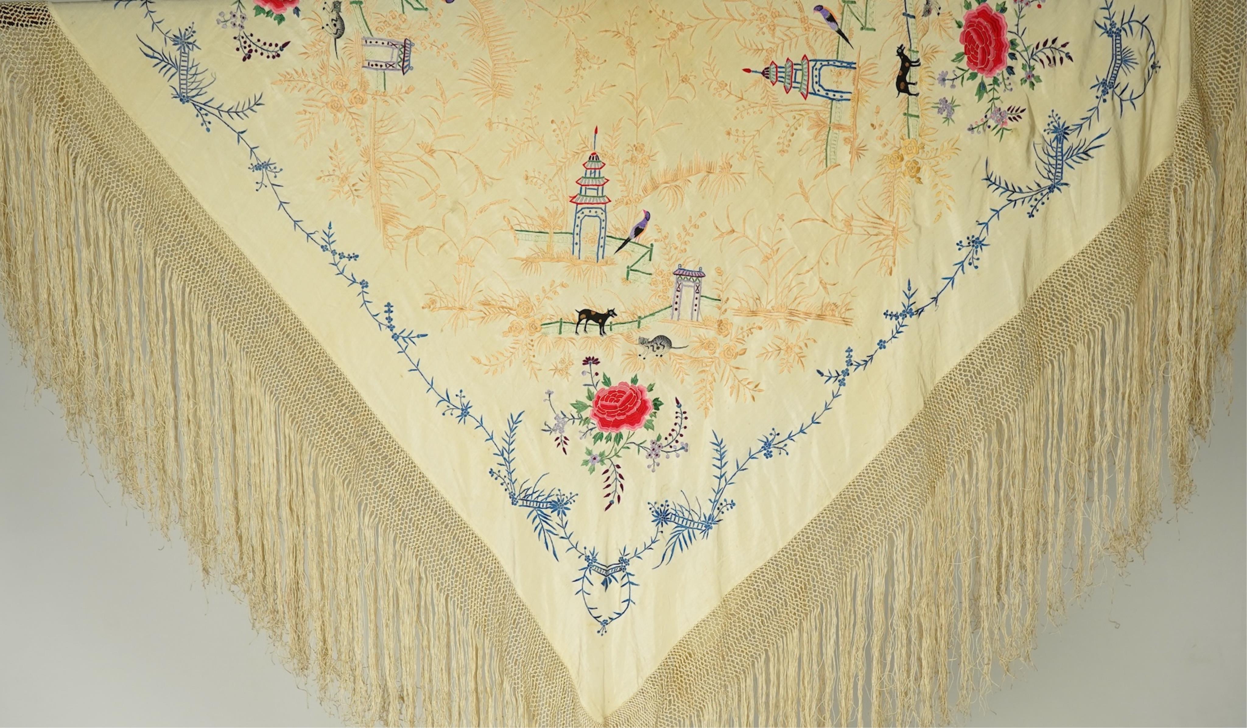 A Chinese cream silk polychrome embroidered shawl with a long fringe. Embroidered with four unusual corner cartouches of Pagoda, flowers, birds, cats and dogs. 132cm x 126cm.